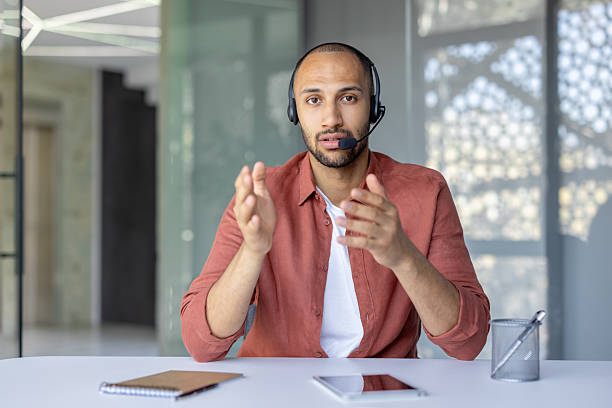 istock Young man with a headset having a video call and gesturing with his hands in an office environment at a white desk. 2210425870