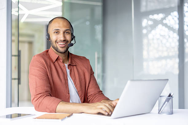 istock Portrait of young online customer support worker. Smiling man looking at camera with headset, using laptop at workplace for video call and support consultation 2210425893