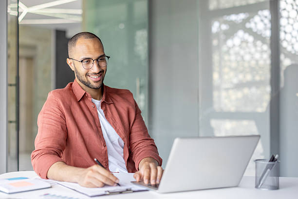 istock A smiling man with glasses works on a laptop while taking notes in a modern office setting, displaying a productive work environment. 2210425908