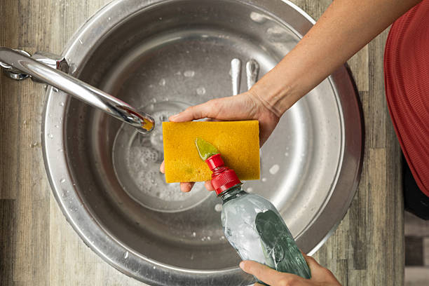 A woman’s hand pours liquid soap onto a sponge over the sink, preparing to clean dirty dishes in a bright and modern kitchen.