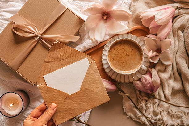 istock A flat lay with a kraft envelope and blank card in hand, next to a wrapped gift box, a coffee cup, blooming magnolia flowers, a lit candle, and soft cozy fabrics 2210455029