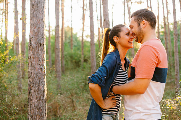 A couple stands in the forest during golden hour, wrapped in each other’s arms and sharing a gentle kiss. The soft backlight from the setting sun creates a warm glow around them, lighting up the trees and casting long shadows. It's a peaceful, romantic moment surrounded by nature’s golden beauty.