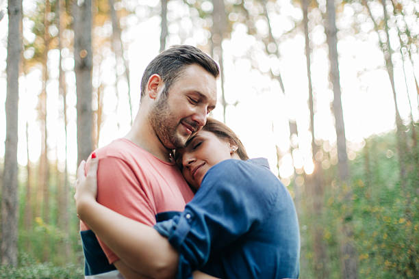 A couple stands in the forest during golden hour, wrapped in each other’s arms and sharing a gentle kiss. The soft backlight from the setting sun creates a warm glow around them, lighting up the trees and casting long shadows. It's a peaceful, romantic moment surrounded by nature’s golden beauty.