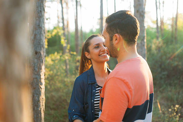 A couple stands in the forest during golden hour, wrapped in each other’s arms and sharing a gentle kiss. The soft backlight from the setting sun creates a warm glow around them, lighting up the trees and casting long shadows. It's a peaceful, romantic moment surrounded by nature’s golden beauty.