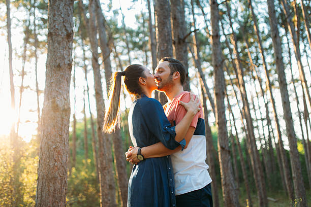 A couple stands in the forest during golden hour, wrapped in each other’s arms and sharing a gentle kiss. The soft backlight from the setting sun creates a warm glow around them, lighting up the trees and casting long shadows. It's a peaceful, romantic moment surrounded by nature’s golden beauty.