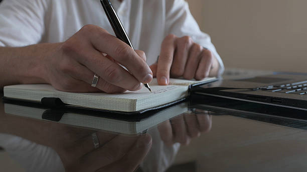 Man Writing Notes at a Desk with a Laptop Nearby