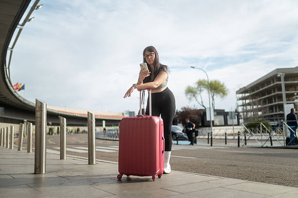 Young woman using smartphone while leaning on suitcase outside airport