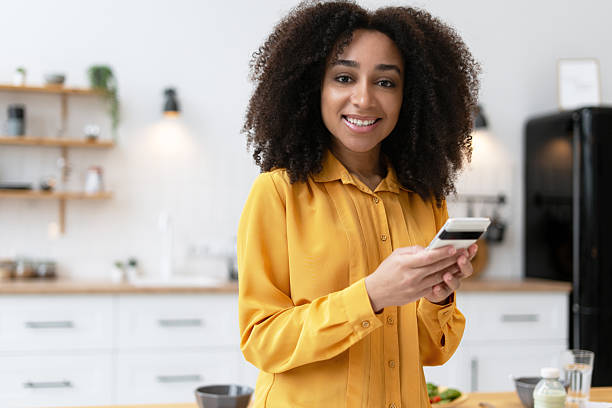 istock Smiling young woman using mobile phone in kitchen 2210495415