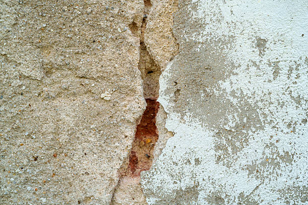 Detailed view of an old exterior wall with visible cracks, peeling white paint, and exposed layers of cement and brick beneath, showing signs of weathering and decay.