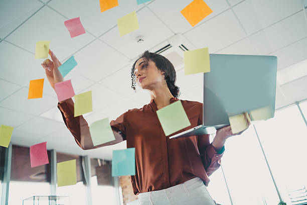 istock Dynamic businesswoman in a modern office using a laptop and organizing tasks with colorful sticky notes on glass walls 2210508331