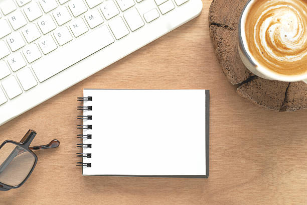 istock Top view, blank notepad with a black spine, spiral binding, computer keyboard, eyeglasses, coffee cup on brown wooden background. Flat lay, space for text or design elements. 2210508579