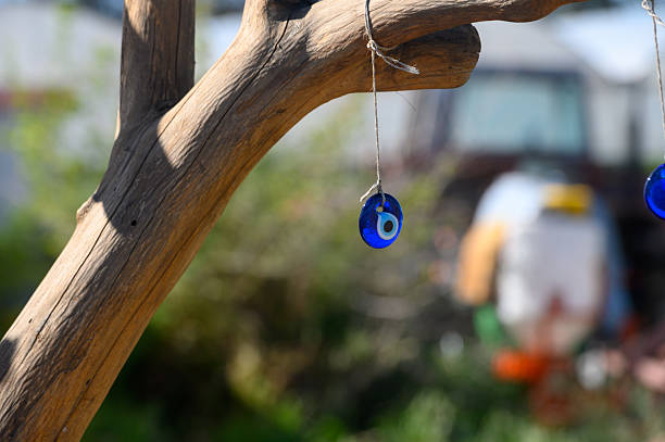 Sunlight filters through a lush garden where blue evil eye charms dangle from a rustic branch. Colorful decorations and greenery create a serene, captivating atmosphere filled with charm.