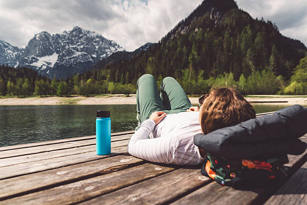 A person relaxing on a wooden dock by a lake, surrounded by mountains and trees. They are lying back with a pillow, enjoying the serene nature. A blue water bottle is nearby.
