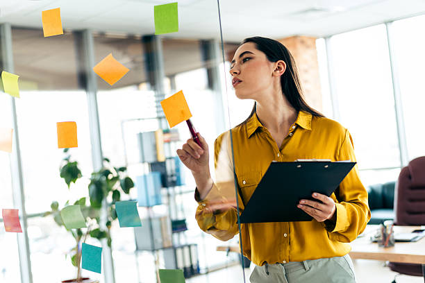 istock Young businesswoman in bright office analyzing ideas on sticky notes while holding a clipboard, focusing on strategy and creativity 2210518398