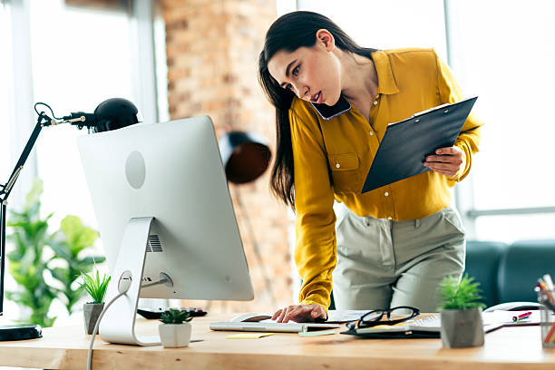 istock Young professional businesswoman working efficiently in a brightly lit modern office environment, multitasking with a phone and documents. 2210518412