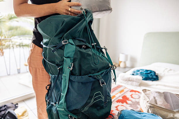 Woman enthusiastically packing a large backpack on a hotel bed. Bright room with a view of palm trees. Ideal for travel, adventure, and packing preparations.