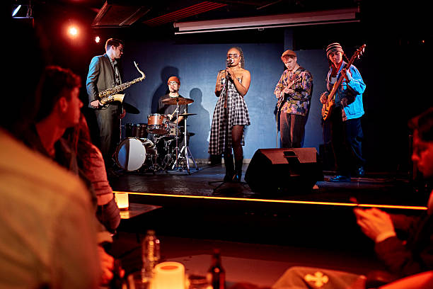 Audience point of view with focus on mature Black chanteuse at microphone, young guitarists, mid adult drummer and saxophonist.