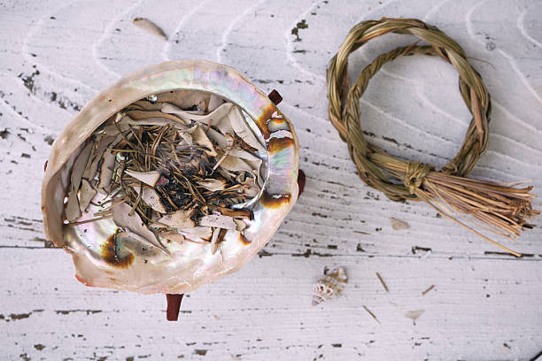 istock Burning sweetgrass (Hierochloe odorata) and white sage (Salvia apiana) in an iridescent abalone shell on an old white wooden table. Overhead view. 2210534725