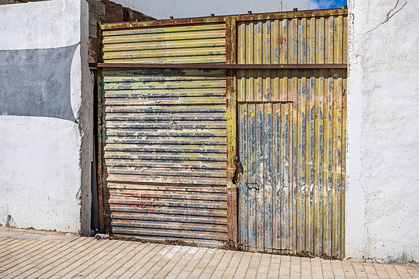 This image features a weathered entrance composed of a rusted corrugated metal gate with a secured door at its center. The faded paint in shades of yellow, green, and blue adds layers of history, while the padlock reinforces the sense of restricted access. The surrounding white concrete walls, marked by patches of wear and graffiti, contrast with the textured metal, highlighting the interplay between decay and security in an urban setting. The picture is taken in Arrecife which is the main city on the Canary Island Lanzarote