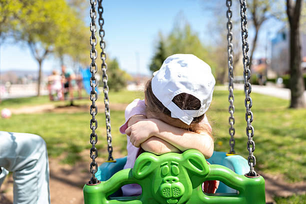A young child is seated on a green swing, leaning forward with arms crossed, resting on the swing's front. The child wears a white cap and is outdoors in a park setting. The sunny background features trees, families, and open play areas, evoking a serene and contemplative moment in a recreational environment.