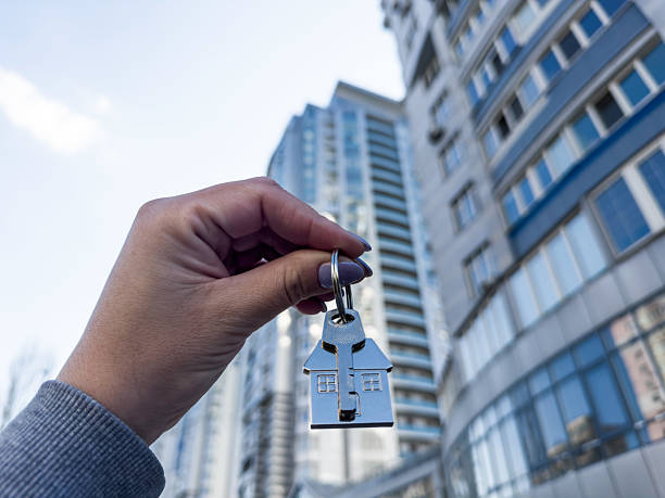 istock A woman's hand holds a house key in plain view in front of a tall, elegant modern building rising in the background. 2210564991