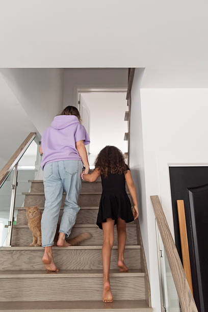 Multiracial mother and daughter going up stairs. Mom is in her forties and daughter is 8 year’s old. Fluffy red cat following them. Vertical full length indoors shot . This is part of a series « day in the life » taken in Quebec, Canada.