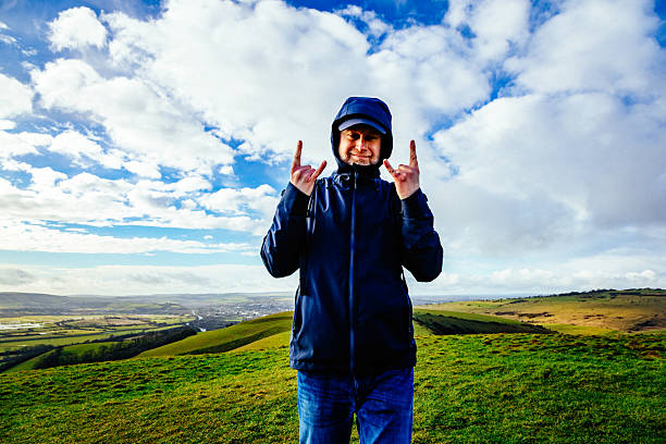 Portrait of a mid adult male hiker outdoors on top of a hill during a hike. The man is wet and exhausted after the storm, and is showing his spirited nature in the face of adversity by doing the 'devil horns' gesture with his hands.