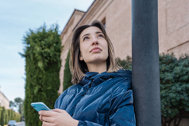 istock Young woman leaning against a lamppost using a smartphone and looking up 2210578320