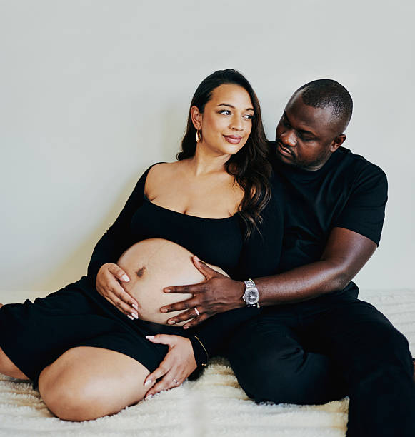 An intimate scene of an expecting couple, lovingly holding the pregnant belly. Both in black attire, the image conveys warmth, love, and anticipation, emphasizing emotional connection and family bonding.