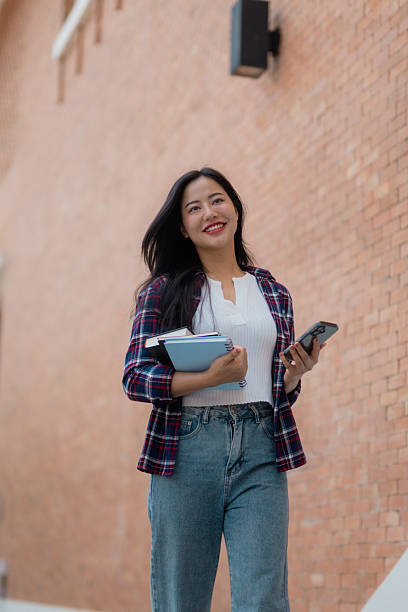 istock Young Asian student walking and looking away while holding books and smartphone 2210591134
