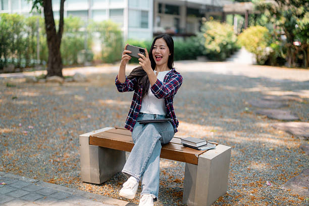 Cheerful young Asian student taking pictures with smartphone sitting on bench in university campus