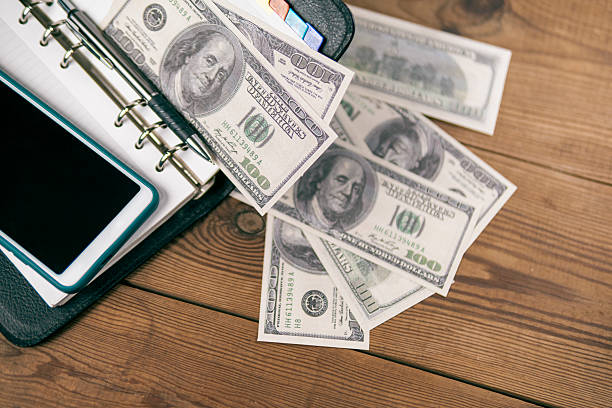 Personal organizer, scattered US Dollar Bills, phone, and pen on a wooden surface.