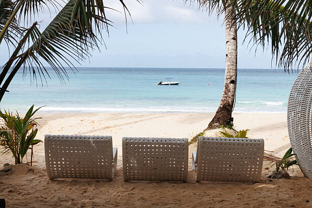 Comfortable lounge chairs are arranged with tropical surroundings on a sandy beach. Gentle waves lap at the shore while boats dot the horizon under a cloudy sky.