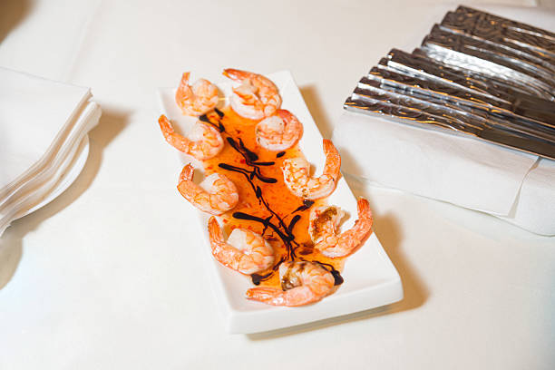Plate of peeled shrimp served with red chili sauce and balsamic drizzle, presented on a white tablecloth with cutlery nearby.