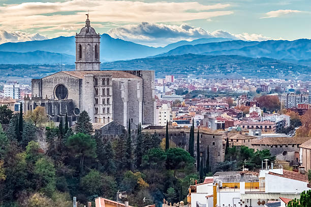 Panoramic view of Girona Cathedral dominating the cityscape with its impressive Gothic architecture against a backdrop of blue mountains, Spain. The historic cathedral sits among cypress trees and stone buildings, overlooking the colorful city, with dramatic cloud formations in the sky above