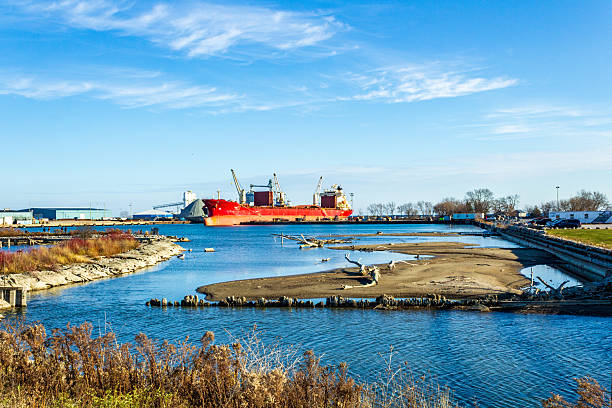 A scenic view of Oshawa Harbour, featuring a bright red cargo ship docked amidst industrial surroundings. Calm waters blend with autumn foliage, complementing the vibrant blue sky and serene atmosphere.