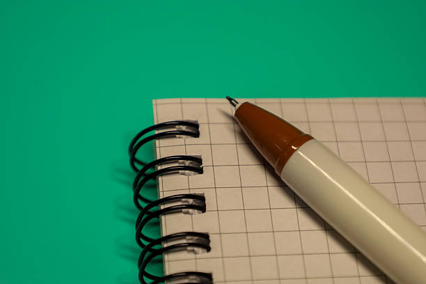 A checkered notebook with a spiral binding and a pen on a bright turquoise background. The pen lies on the open page of the notebook, ready for use. A simple and minimalist image, ideal for illustrating concepts of writing, learning, planning, notes, and creativity.