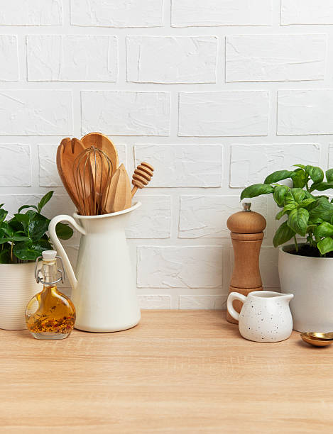 Wooden kitchen utensils, fresh basil, oil, and condiments arranged on a wooden countertop against a white brick wall, creating a cozy culinary scene