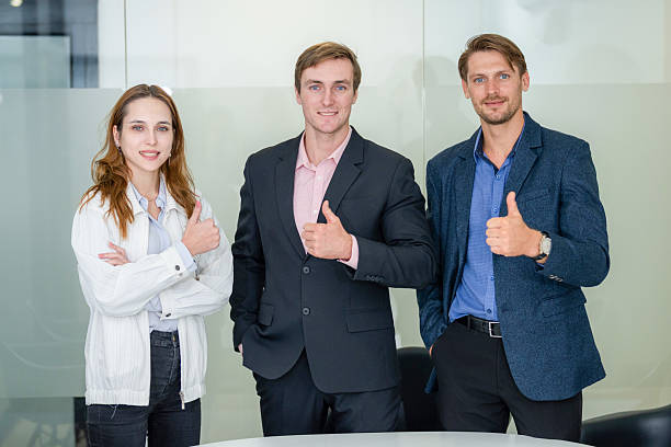 Three professionals stand confidently in a contemporary office, giving thumbs up gestures. The setting is bright and well-lit, suggesting a positive atmosphere.