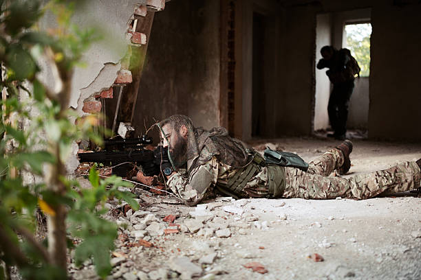 A soldier dressed in camouflage and tactical gear aiming a weapon inside a dimly lit building, with rays of sunlight streaming through a window.