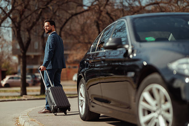 Rear view of a businessman in formal attire pulling a suitcase, walking away from his parked car, heading toward his destination in an urban area.