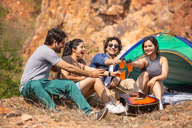 Cheerful young friends toasting drinks while sitting with guitar outside tent at campsite during weekend getaway
