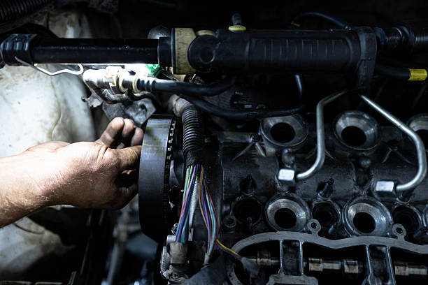 A mechanic's hand works on the timing belt area of a partially disassembled engine inside a vehicle, illuminated by a work light.