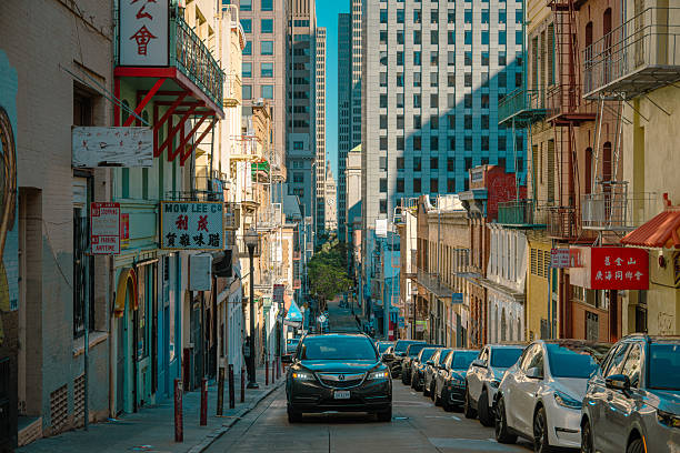 A cinematic view down a narrow and steep street in San Francisco’s Chinatown. The street is lined with parked cars, traditional Chinese storefronts, and signage, giving way to modern downtown skyscrapers in the distance. The juxtaposition of cultural heritage and urban development highlights the city’s layered identity and diversity.