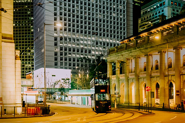 A city street scene features a tram passing by tall buildings under a warm evening sky with lights glowing.