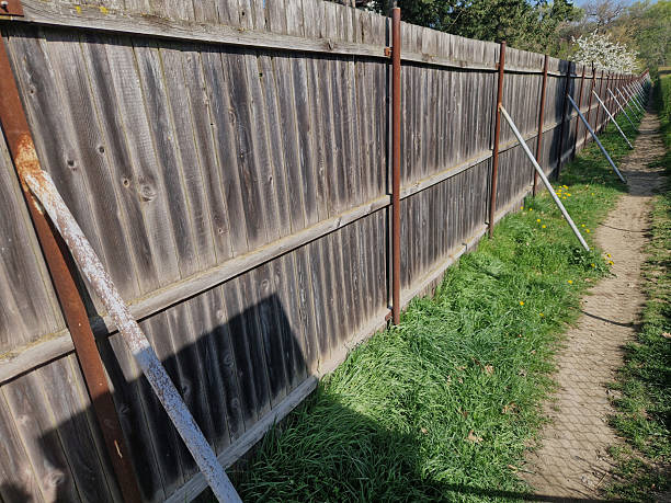 A plank fence without gaps is prone to being knocked over in strong winds. Pipe struts support the heavy fence from the sides. A path along
