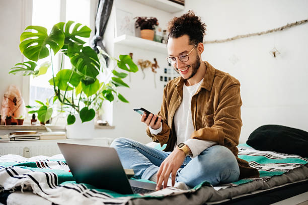 istock Male student enjoying remote study session with Laptop while sitting on bedsmiling at phone screen 2210683428
