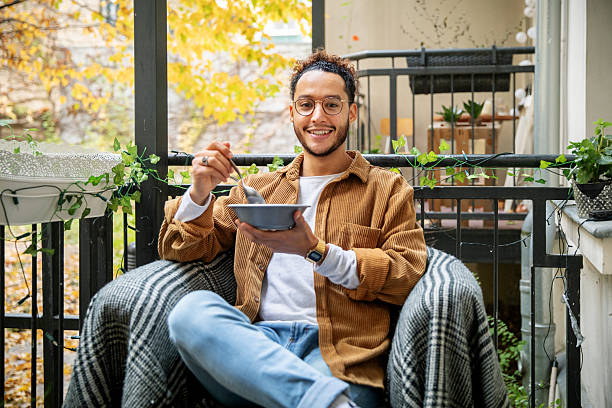 A young man sits comfortably on a blanket-draped chair on a small balcony, eating cereal from a bowl. Surrounded by autumn colors and decorated with potted plants, the setting is serene and perfect for a peaceful start to the day.