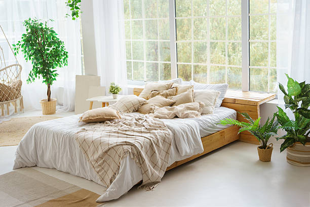 Comfortable bed with lots of soft decorative beige pillows in light sunny bedroom with green plants and large window. Interior design of living room in boho style