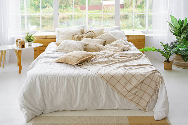 istock Comfortable bed with lots of soft decorative beige pillows in light sunny bedroom with large window 2210695949